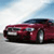 A red sports car with a black roof is positioned on a light gray surface under a blue sky with white clouds.