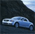 A light blue sedan with dark blue accents is positioned on a dark road under a dark sky, viewed from a low angle with a light blue sky in the background.