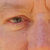 A pixelated close-up of a human eye with brown and gray tones, showing eyelashes and partial eyelid details against a blurred peach-toned background.