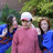 Three people pose outdoors with green foliage background; central figure wears pink shirt and white cap, flanked by two others in blue tops.