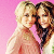 Two people with light and dark hair stand close together against a solid pink background, both smiling.