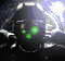 A dark, helmeted figure with glowing green eyes and a small green dot on its chest, set against a bright, blurred background with light streaks.