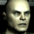 Pixelated close-up of a bald human face with neutral expression, dark eyes, and visible ear against black background.