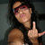 A person with long dark hair and red-tinted glasses makes a middle finger gesture with their right hand against a dark background.