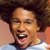 Pixelated close-up of a person with dark curly hair, light skin tone, and an open mouth showing teeth against a blurred background.