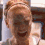 A pixelated image of a person with light brown hair in a bun, wearing a light-colored top, with visible background elements including vertical structures and a dark rectangular shape.