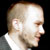 Static 50x50 pixel icon showing a close-up profile of a bald man with light skin, wearing a white shirt and gray jacket against black background.