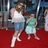 Two figures stand on a red carpet; one wears a white sleeveless top and blue pants, the other a green and white dress, with a blurred background of vertical elements.
