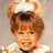 Pixelated image of a child with brown hair styled in two buns, wearing white clothing and smiling against a light background.