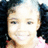 A young girl with dark curly hair in pigtails, wearing a pink top and smiling broadly against a black background.