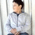 A person in a light gray zip-up jacket with dark trim sits against vertical white , hands clasped over the lap
