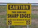 Yellow caution sign with black text warning about sharp edges, displayed outdoors on a grassy field background.