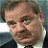 Pixelated close-up of a person with short brown hair, light skin, and a mustache, wearing a dark suit jacket over a white shirt, against a blurred background.