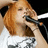 A pixelated close-up of a person with light brown hair holding a microphone near their mouth, wearing a white top with dark patterns.