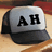 A pixelated baseball cap with gray front panel and black brim, displaying large black letters 'AH' on the front against a blurred background.