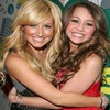 Two women embrace in a close-up pose, one with long blonde hair in a dark sleeveless top, the other with long brown hair in a red sleeveless dress, both smiling broadly.