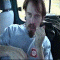 A sequence of animated frames showing a person in a light blue shirt with red logo, seated inside a vehicle, performing repeated hand-to-face gestures against a blurred background.