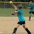 Pixelated image of a person in a teal shirt and black shorts throwing a yellow ball on a sandy field with green grass in the background.