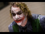 Static pixelated image of a character with white face paint, red lips, and green vest over dark shirt against blurred background.