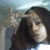A person with long dark hair makes a peace sign with their right hand while wearing a white shirt against a light blue background.