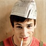 Static image of a person wearing a newspaper hat, red shirt, and necklace with a yellow object in mouth against beige background.