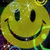 A pixelated yellow smiley face with black oval eyes and a wide curved mouth, set against a blurred multicolored background.