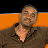 Pixelated image of a person with short hair wearing a dark jacket over a light shirt, seated against an orange and black background.