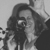 Static grayscale image of a person with long hair holding an object near their face, set against a plain background.