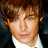 Pixelated close-up of a person with light brown hair, wearing a white shirt and dark jacket, against a dark background.