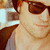 Pixelated close-up of a person with brown hair wearing dark sunglasses and a light-colored top, holding an object near their face.