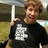 A pixelated image of a person in a black t-shirt with white text, holding a white pole, next to green pixelated text on black background.