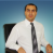 Pixelated image of a person in business attire against a blue gradient background, wearing white shirt and dark tie with visible chair edge.