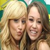 Two women with long hair pose closely together; one has blonde hair and the other brown hair, both with neutral to smiling expressions.