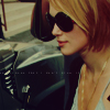 A close-up of a person with blonde hair wearing dark sunglasses and a light-colored top, positioned next to a dark vehicle with visible headlights.