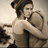 Pixelated image of two figures in a close embrace, one with dark hair tied up, the other with light brown hair, both in neutral-toned clothing against a muted background.