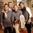 Four individuals in casual attire pose closely together with arms around each other against a blurred background.