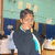 Pixelated image of a person with short dark hair wearing a dark jacket over a light shirt, standing in front of a blue and beige background with blurred shapes.