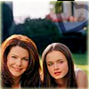 Two women with long brown hair pose outdoors; one smiles broadly while the other has a neutral expression, set against a blurred background of greenery and a blue vehicle.