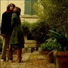 Two figures embrace in a garden setting with a building facade and greenery in the background.