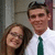 Two people in formal attire pose together; the right person wears a white shirt and green tie, while the left person has long brown hair and glasses.