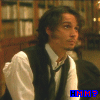 A person in a white shirt and dark vest sits before bookshelves, with blue text 'HMM?' at the bottom right.