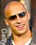 Pixelated close-up of a person with light skin, short blonde hair, dark sunglasses, and a slight smile wearing a white shirt.