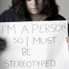 A person with shoulder-length brown hair holds a white sign with handwritten text against a plain light background.