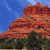 Pixelated landscape showing a large red rock formation under a blue sky with scattered clouds, with green vegetation at the base.