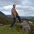 Pixelated static image of a person standing outdoors on grassy terrain with a body of water and sky in the background.