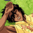 Pixelated image of a person with dark curly hair lying on green grass, wearing a yellow shirt with red text, hand on forehead, next to brown object.