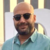 Pixelated image of a bald man with dark sunglasses, brown beard, and light blue collared shirt against a striped background.