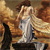 A pixelated figure in a white gown stands on a dark platform with a brown-haired head turned to the right, set against a warm-toned background of orange and brown hues.