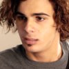 A close-up portrait of a person with curly brown hair wearing a gray shirt against a light background.
