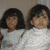 Two children in white shirts with colorful graphics sit side-by-side against a light background, one child's face partially obscured.